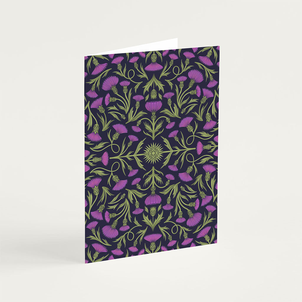 The Heritage Thistle Greetings Card stands upright and features a symmetrical design of green stems and purple thistle flowers on a dark background.