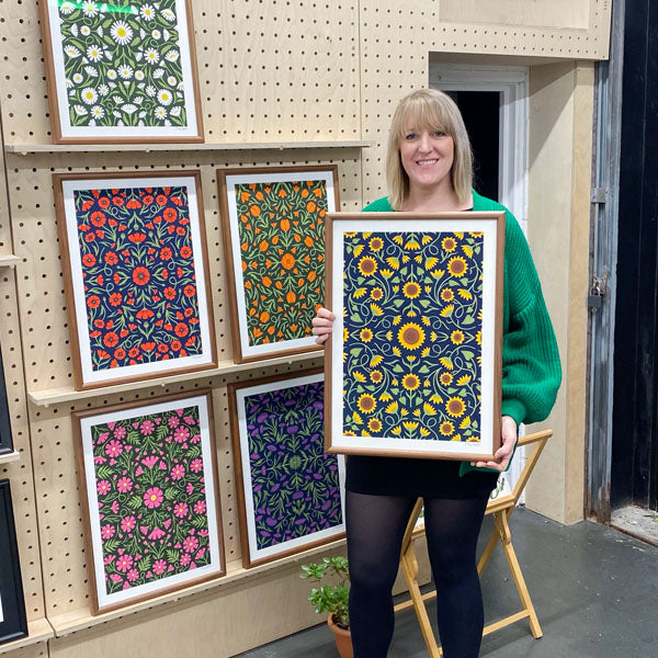 A woman stands indoors holding a framed floral artwork, with several other colorful floral prints displayed on a pegboard wall behind her.