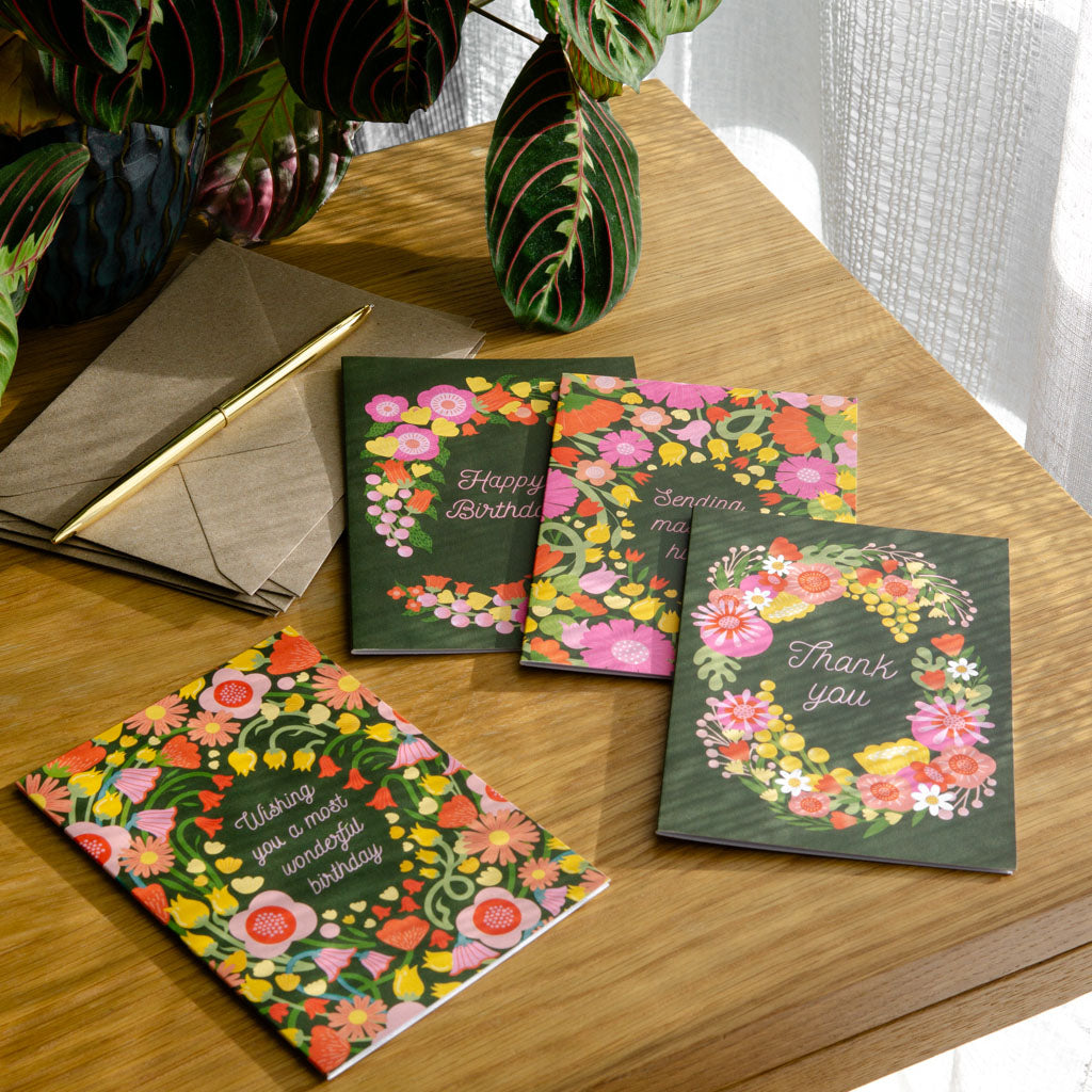 The Vibrant Florals Greetings Card Set includes four cards with heartfelt messages—birthday wishes, thank you, happy birthday, and sending hugs—all on dark green floral backgrounds for any occasion.