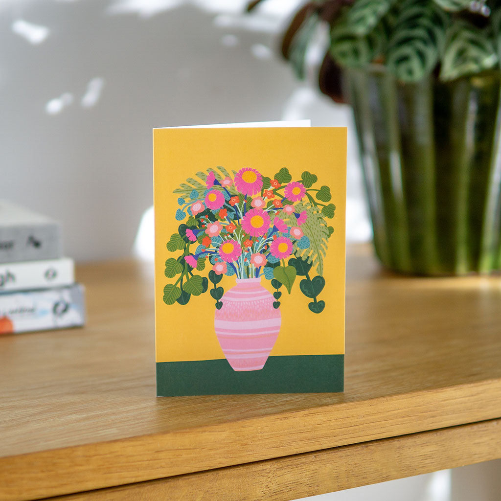 The Flowers Vase Greetings Card features a pink vase with colourful blooms and green leaves on a yellow-green background, making it perfect for birthdays or any occasion.