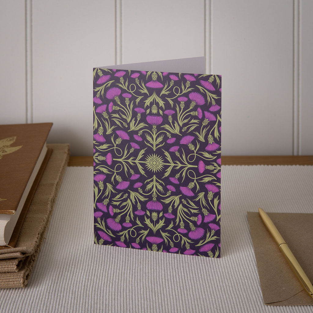 The Heritage Thistle Greetings Card stands upright and features a symmetrical design of green stems and purple thistle flowers on a dark background.