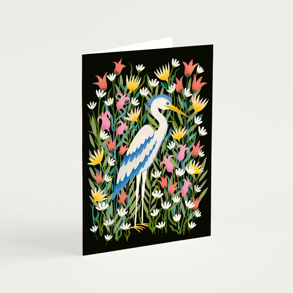 The Heron Greetings Card features a white and blue heron standing among colourful flowers on a black background—ideal for birthdays or as a thoughtful Mothers Day card.