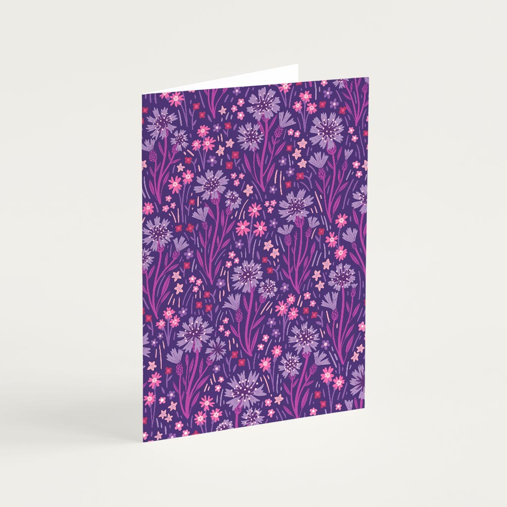 The Indigo Wildflowers Greetings Card stands upright, showcasing a decorative purple background with pink, lavender, and indigo wildflowers and foliage.