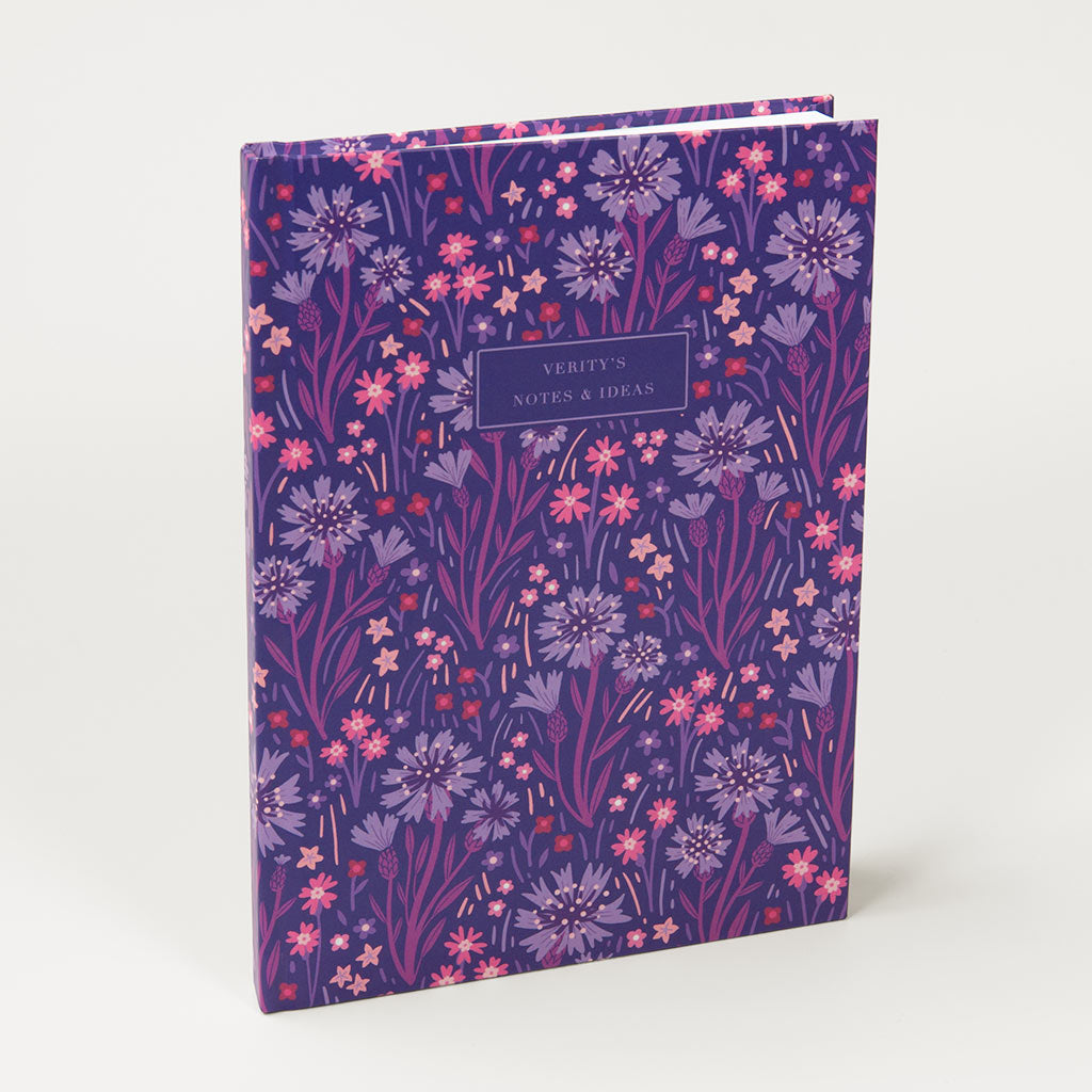 The Indigo Wildflowers Personalised Notebook is a purple hardcover journal featuring pink and lavender British wildflower illustrations, with a customised front label.