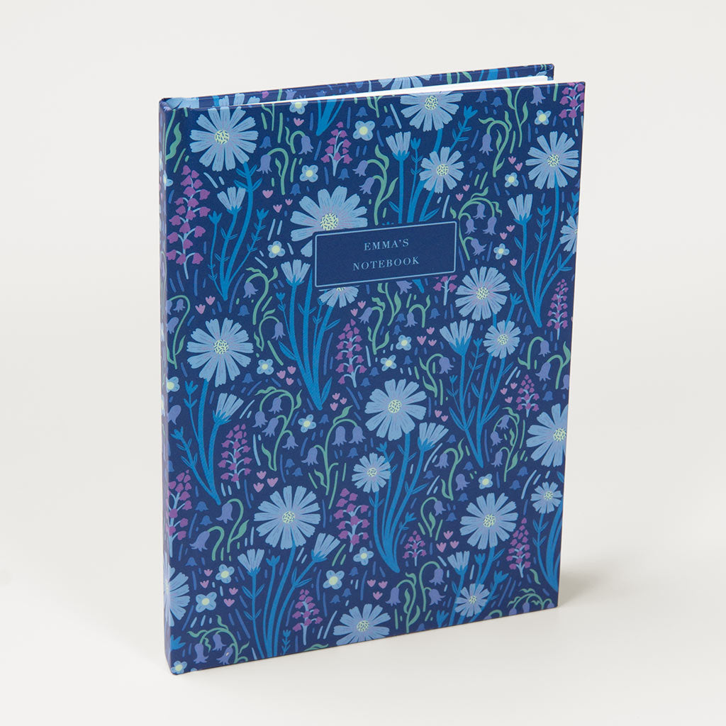 The Navy Blue Wildflowers Personalised Notebook, featuring a blue floral pattern inspired by British wildflowers and a custom label section, stands upright against a white background.