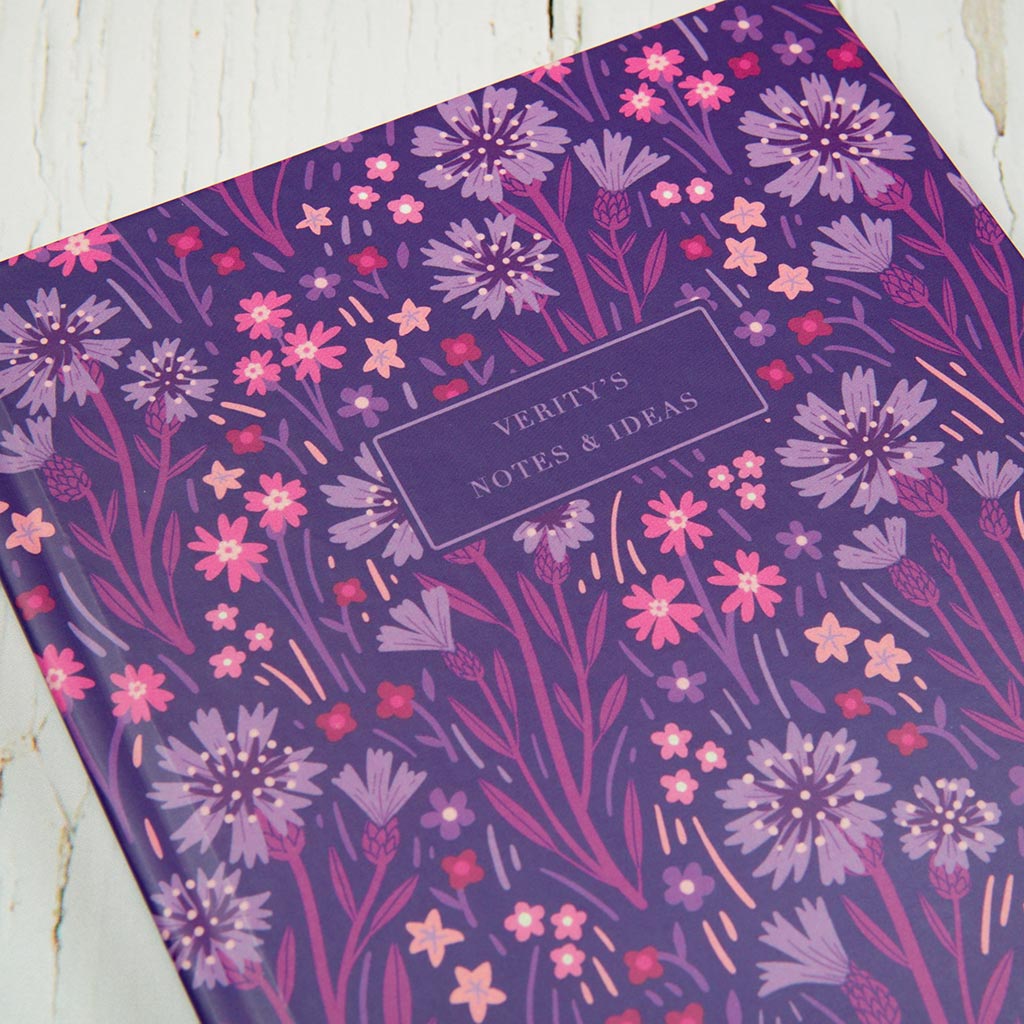 The Indigo Wildflowers Personalised Notebook features a pink and purple floral design inspired by British wildflowers, with a personalised journal label in the centre, and is displayed on a white wooden surface.