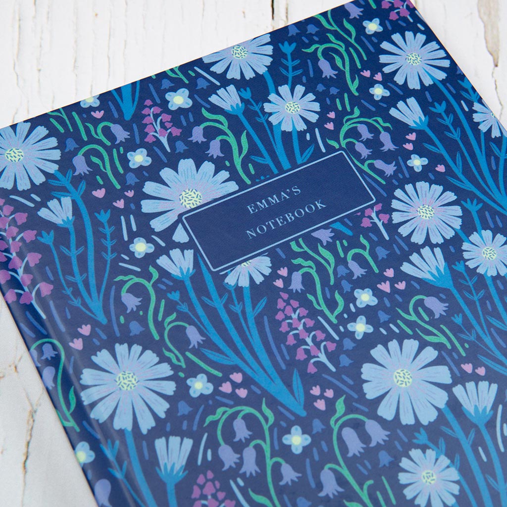 The Navy Blue Wildflowers Personalised Notebook, featuring a blue floral pattern inspired by British wildflowers and a custom label section, stands upright against a white background.