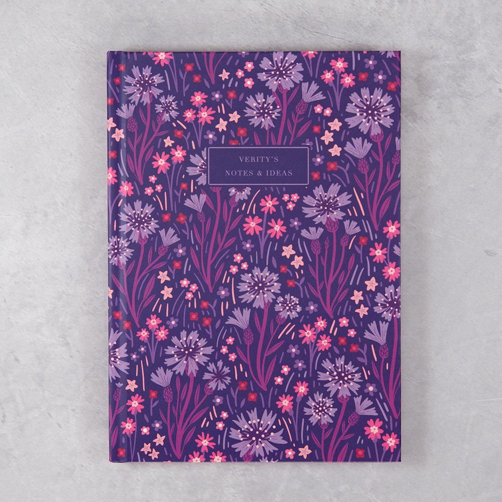 The Indigo Wildflowers Personalised Notebook features a purple cover with British wildflower illustrations and a personalised text label, displayed on a light grey surface.