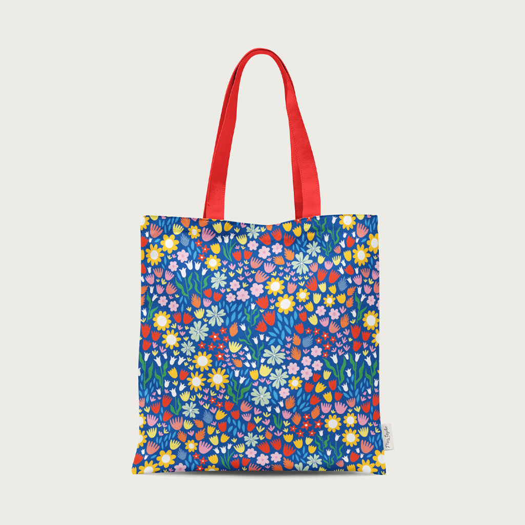 The UK-made Spring Bloom Tote Bag, crafted from organic cotton canvas, features red handles and a vibrant floral pattern on a blue background.