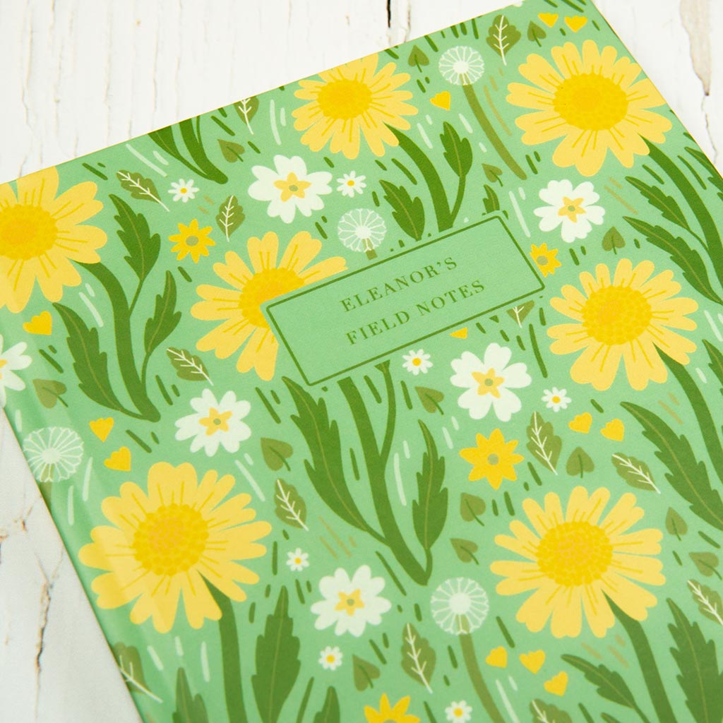 The Green Wildflowers Personalised Notebook features charming British wildflower illustrations, a yellow and white daisy pattern, and a customisable text label on the cover.