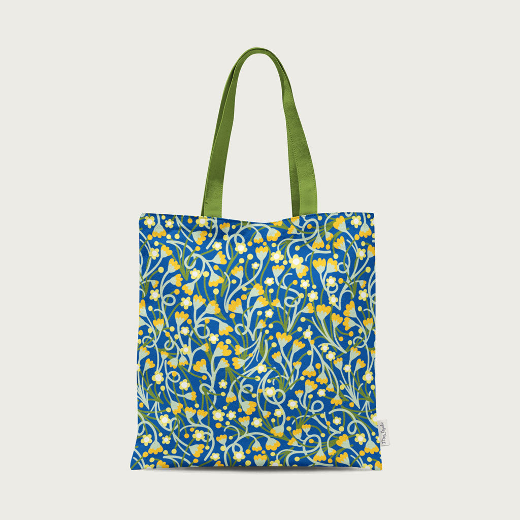 The Wildflower Meadow Tote Bag is crafted from organic cotton canvas with green handles and a blue background decorated with cheerful yellow and white flowers.